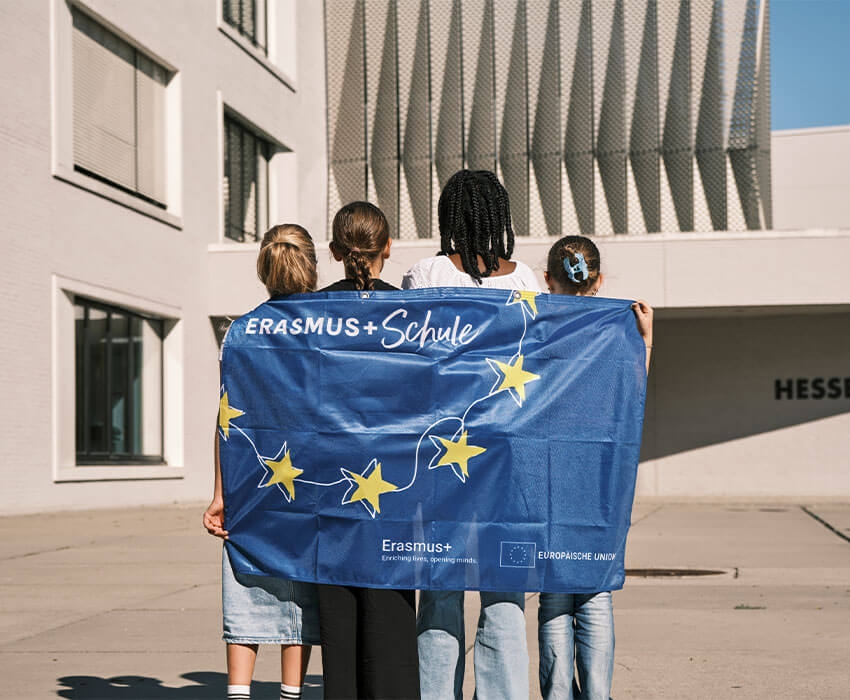 Kinder stehen mit dem Rücken zur Kamera und haben die Erasmus+ Flagge an ihrem Rücken und schauen dabei zur Schule