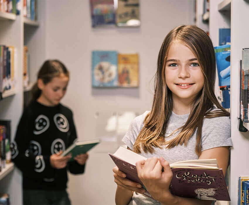 Zwei Mädchen stehen zwischen zwei Bücherregalen und halten ein Buch in der Hand dabei lächeln sie in die Kamera
